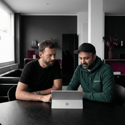 Josip trägt ein schwarzes T-Shirt und sitzt mit Sascha, der eine Cappy und einen grünen Hoodie trägt, an einem Tisch und schaut mit ihm auf ein Tablet