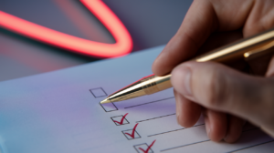 Nahaufnahme einer Hand, die mit einem goldenen Kugelschreiber ein Kontrollkästchen auf einer Checkliste markiert. Mehrere Kästchen darunter sind bereits mit roten Häkchen versehen.